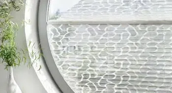 Rundes Fenster mit weißem, gemustertem Plissee und Zimmerpflanze.